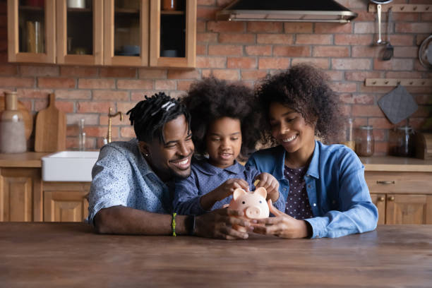 A smiling young African American family with a daughter saves money in piggybank feel provident economical about finances. Happy biracial parents with ethnic child make financial investment for future.