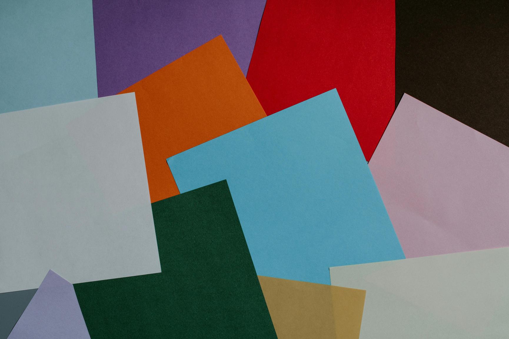 Overlapping sheets of colourful paper in red, blue, green, orange, purple, pink and white arranged in a flat lay composition.