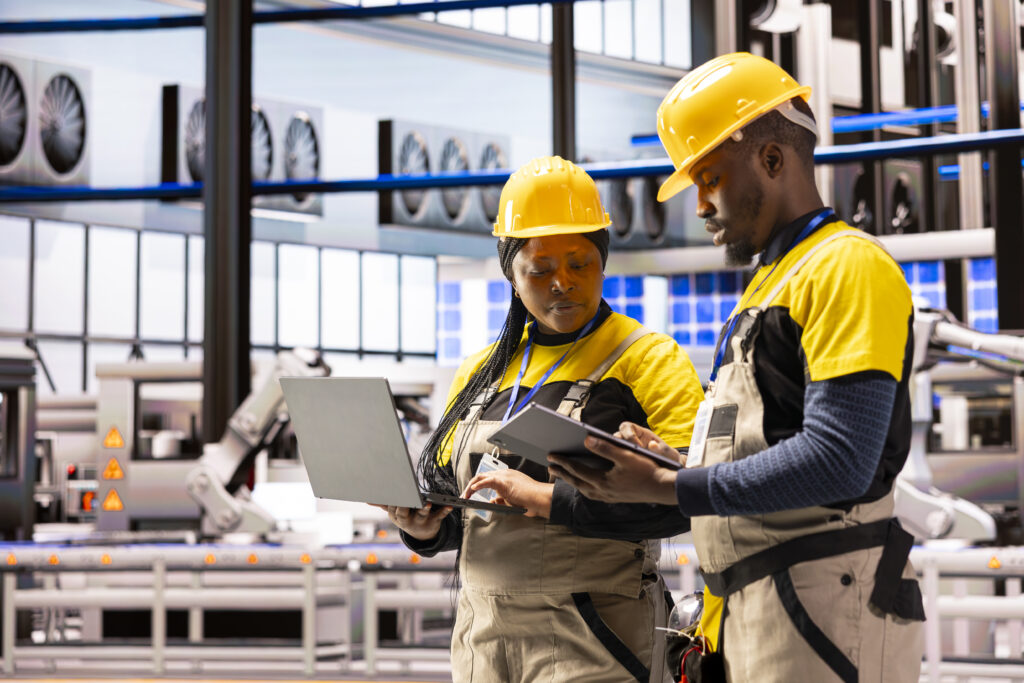 Engineering team in smart manufacturing plant using tablet and laptop for quality control, ensuring smooth industrial automation. Technicians manage assembly lines and conveyor belts in smart factory.